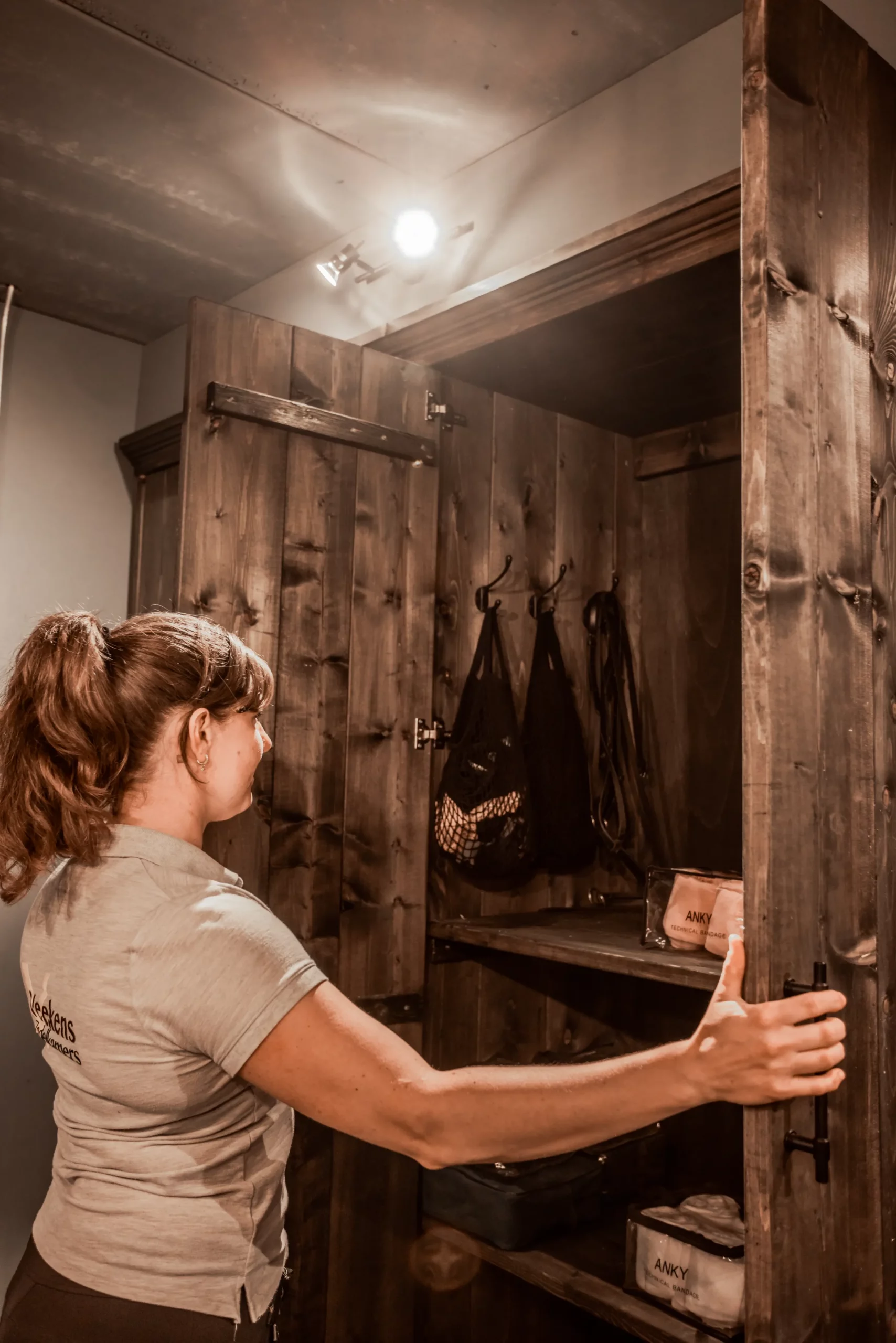 Tackroom. Vrouw opent donker houten zadelkast, kleding, haken met netjes.