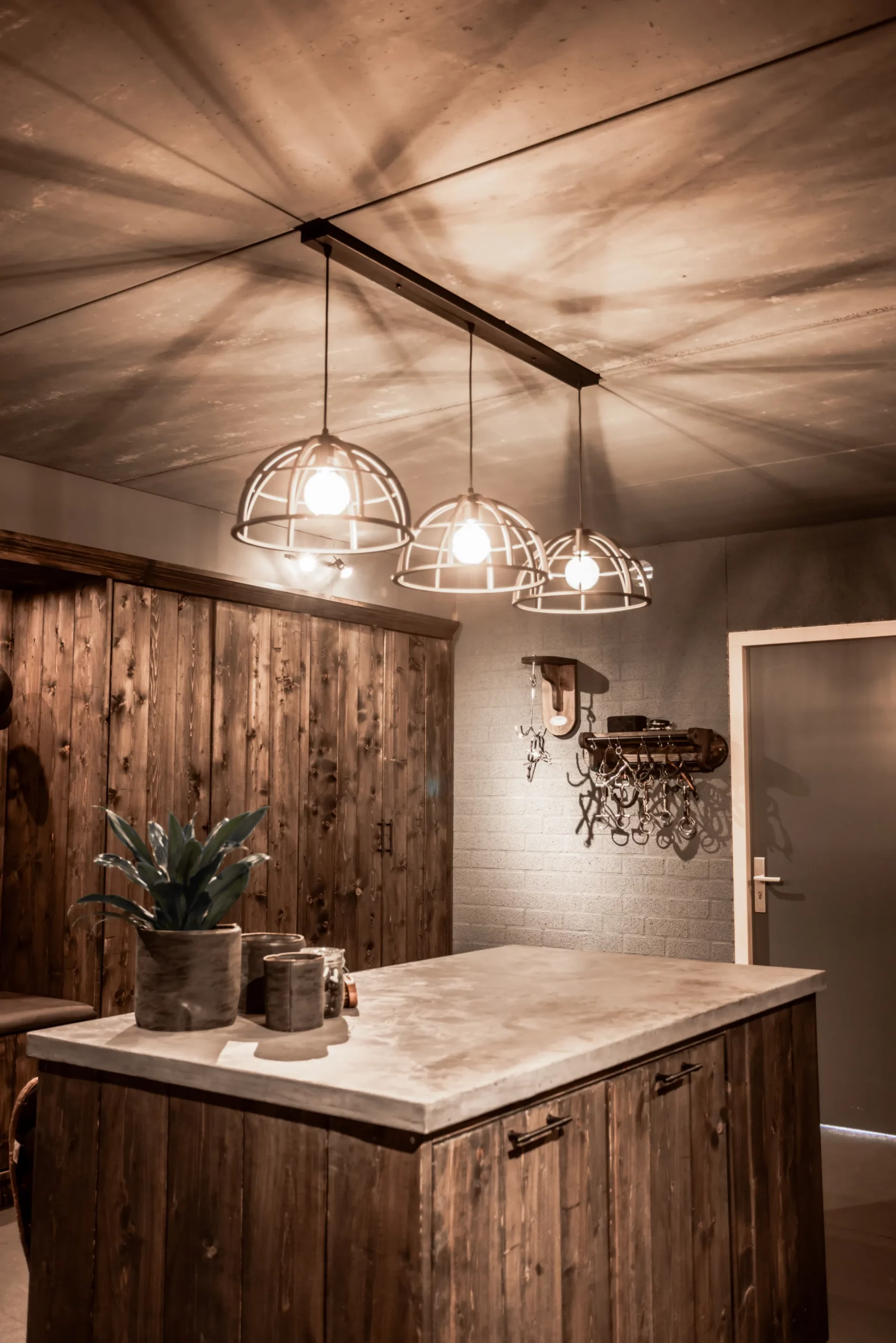 Tackroom. Donker houten zadelkast, grijze muur en deur. Kastmeubeleiland, ijzeren lampen, poetshaak met bit, bittenrek met bitten, decoratie.