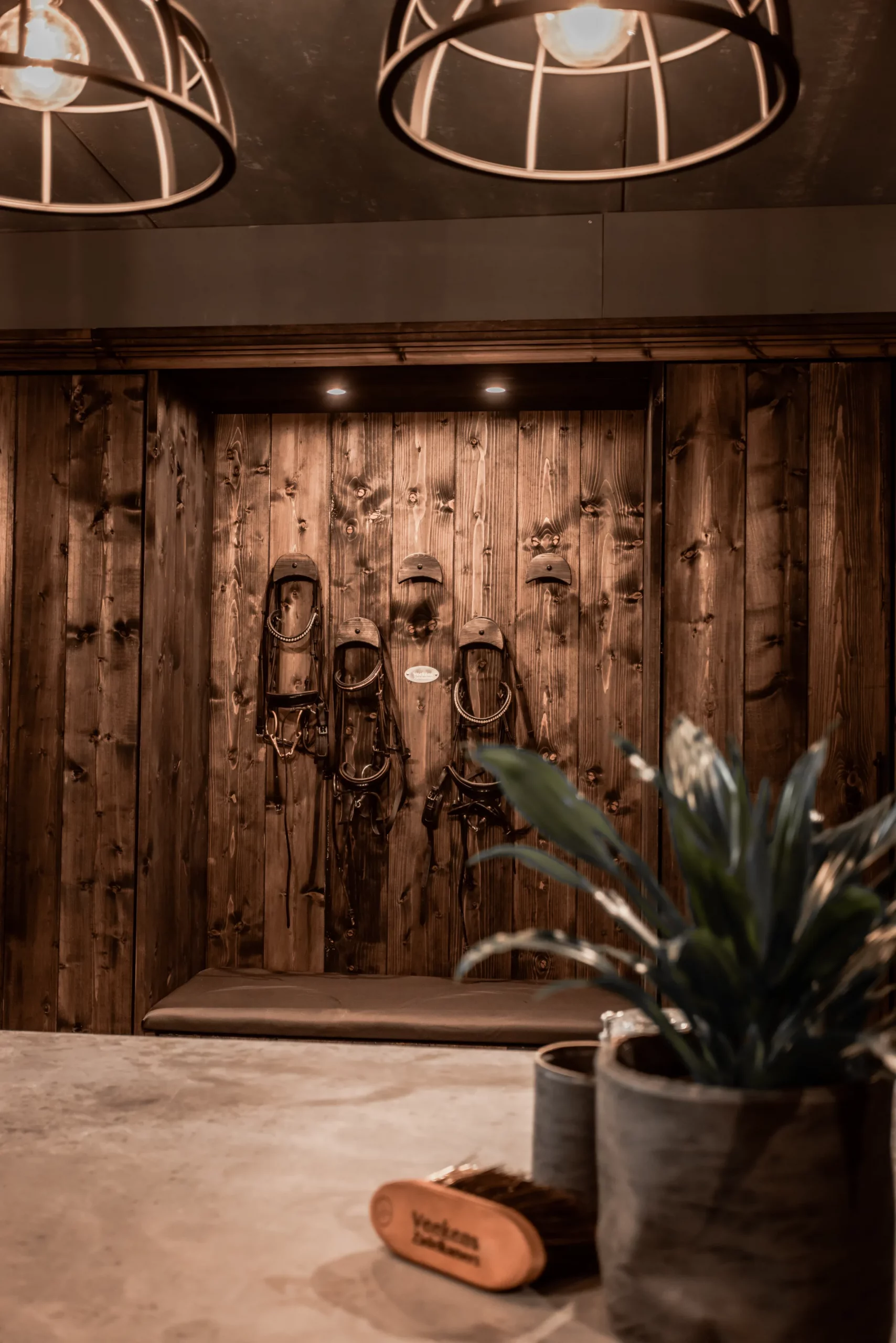 Zadelkamer. Sfeerbeeld donker houten zadelkast vanaf meubeleiland. Hoofdstel haken met hoofdstellen, plant, ijzeren lamp.