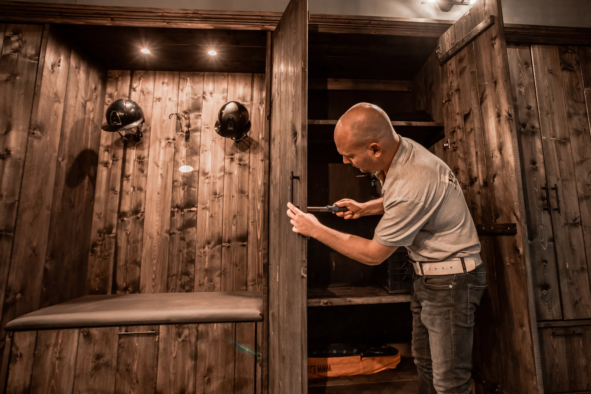 Veekens zadelkamers medewerker instaleert donker houten zadelkast, laarzenmeubel, capholders met helmen.