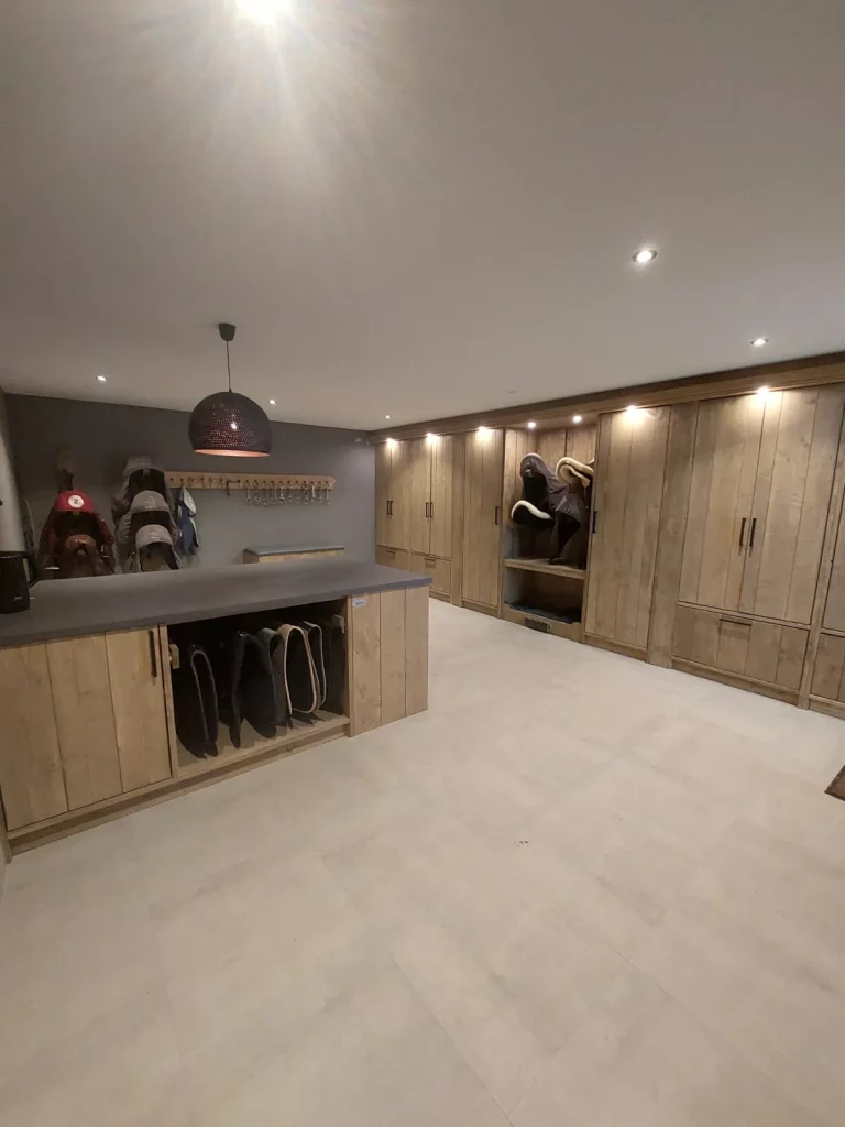 Wit-grijze tack room. Meubeleiland van kastjes en paarden deken steunen. Hoofdstel haken met paarden hoofdstellen. Laarzenmeubel, zadelkastjes met deur en zadelsteunen.