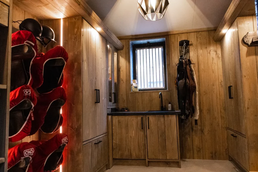 Donkere houten tackroom grijs met luxe ledverlichting. Zadelsteunen met zadels, capholders met helmen, vitrine kast, gesloten kasten, hoofdstel haak, poetshaak met paardrijd materiaal, wastafel, ijzeren raam.