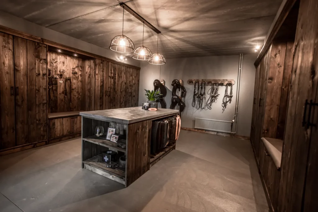 Luxe tackroom zwart hout. Houten gesloten zadelkasten, inhammen met laarzenmeubels, hoofdstelplanken met hoofdstellen, zadelsteunen met zadels, meubeleiland met uitschuifbaar dekenrek, verwarming, ijzeren lampen, plant, decoratie.