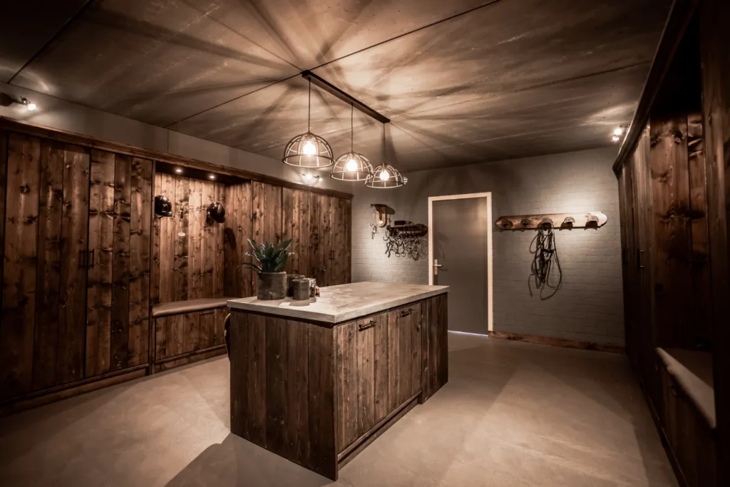 Luxe tackroom zwart houten. Meubeleiland met gesloten kastjes, hoge gesloten kasten, grijze deur en muur hoofdstel haken met hoofdstel, poetshaak, bittenrek met bitten, laarzenmeubels, capholders met helmen, ijzeren lampen.